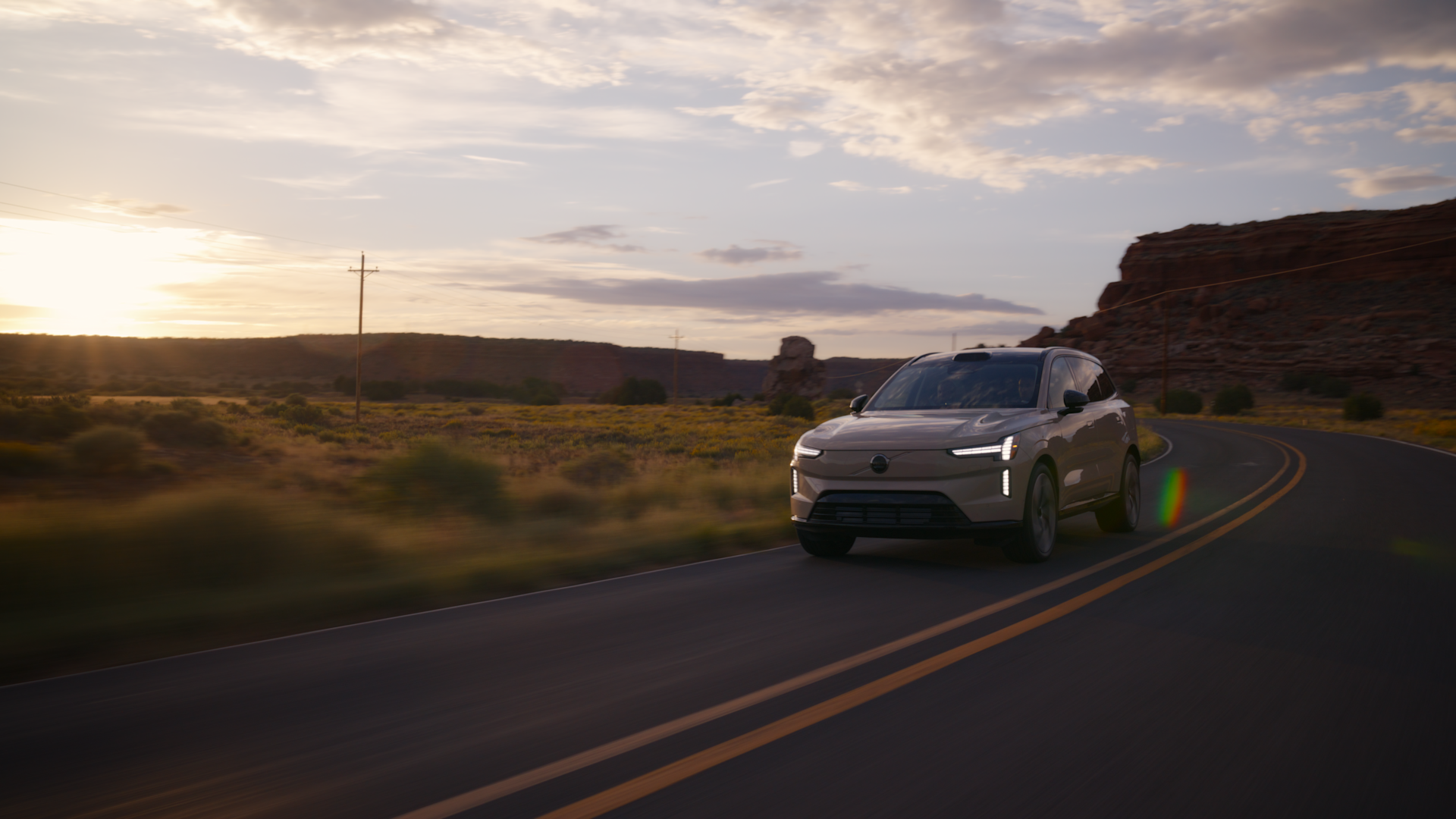 A white SUV drives down a winding desert road at sunset, with rocky formations and open landscape in the background under a partly cloudy sky.