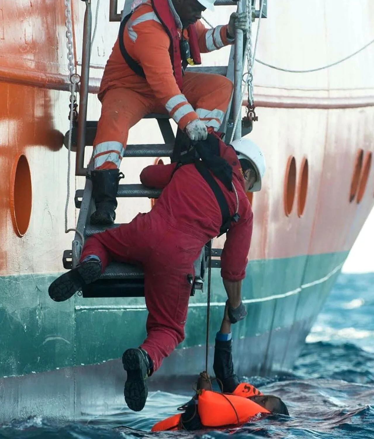 Two crew members in safety gear help rescue a person in an orange life vest from the ocean beside a ship, using a rope ladder and teamwork to pull them out of the water.