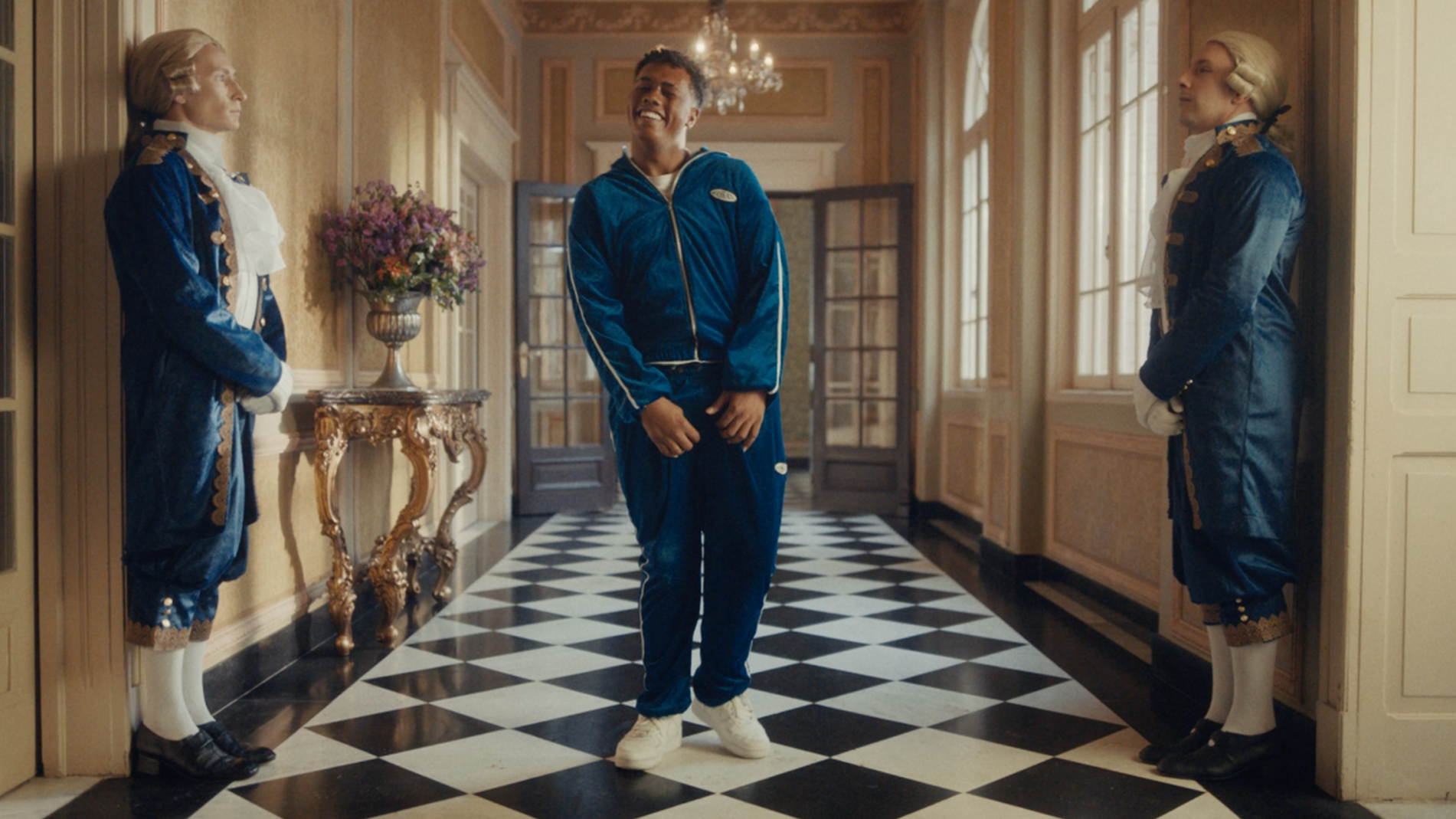 A man in a blue tracksuit stands smiling in a grand hallway with checkered floors, flanked by two people in 18th-century-style blue uniforms, standing by the walls. A chandelier and large windows add elegance to the scene.