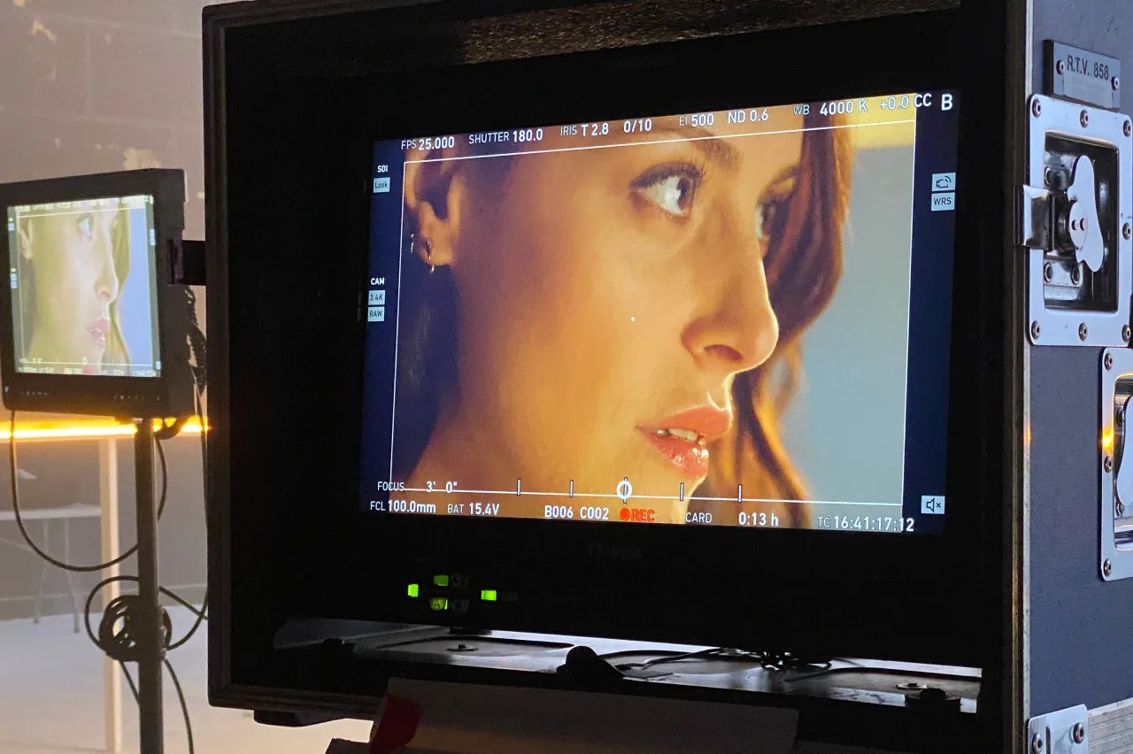 A close-up of a womans face appears on a monitor during a film shoot in a studio, with film equipment and another monitor in the background showing the same image.