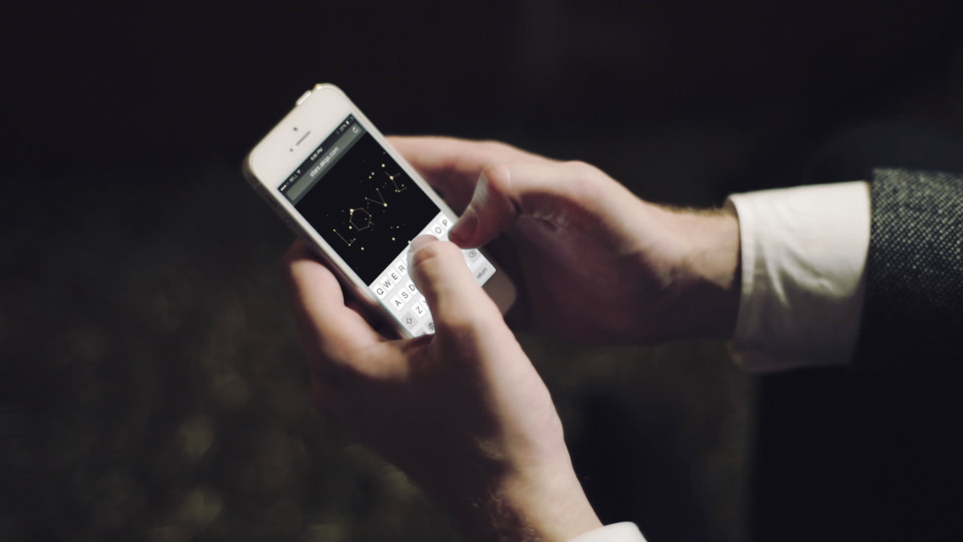 A person holding a smartphone and typing a message. The screen displays the word LOVE written in a stylized font on a black background. The person is wearing a white shirt and a grey jacket.