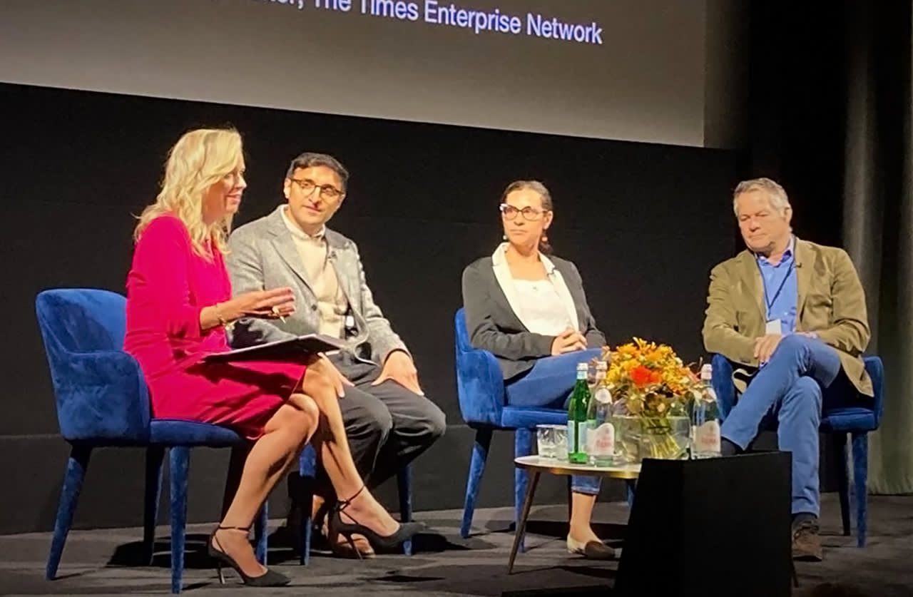 Four people sit on stage in blue chairs, engaged in a panel discussion. One woman speaks while holding a microphone, with three panelists listening. A table with flowers and water bottles is in front of them.