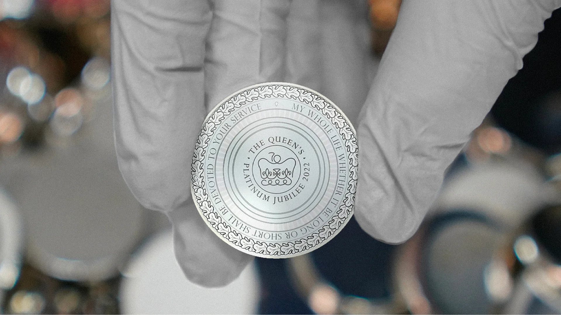 A gloved hand holds a silver coin featuring the inscription The Queens Platinum Jubilee 2022 surrounded by a decorative border and additional text around the coins edge.