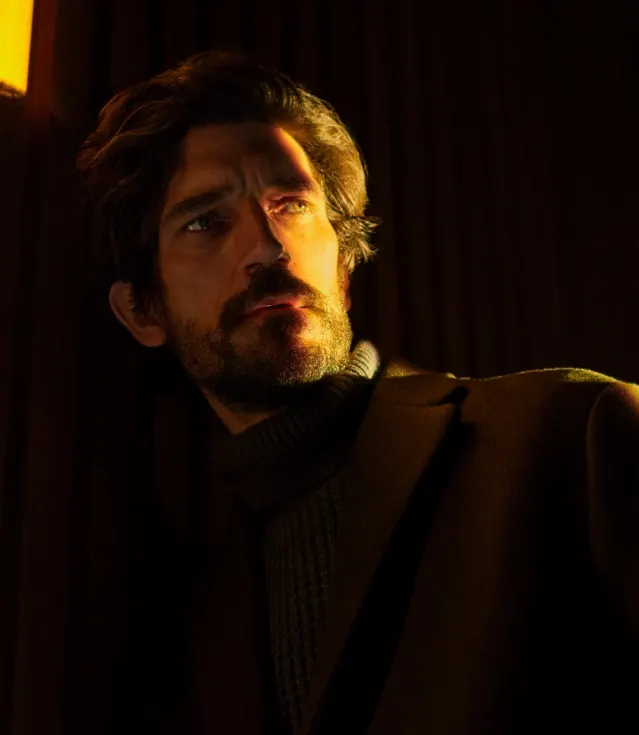A man with wavy dark hair and a beard stands in dramatic lighting, casting shadows on his face. He wears a dark coat over a ribbed turtleneck sweater and looks pensively into the distance.