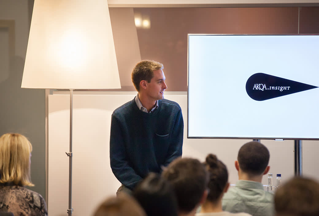 A man in a blue sweater sits in front of a group, facing sideways toward a screen displaying the text AKQA insight. The audience listens attentively in a modern, well-lit room.