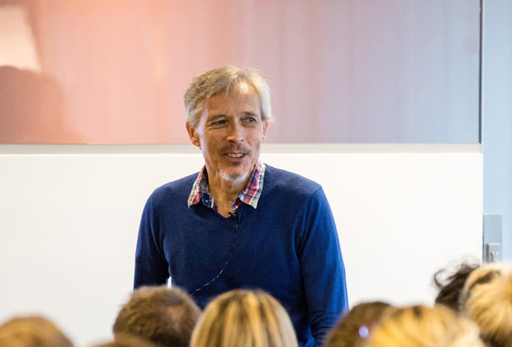 A middle-aged man with gray hair, wearing a blue sweater over a plaid shirt, stands and smiles while speaking to a seated audience in a brightly lit room.