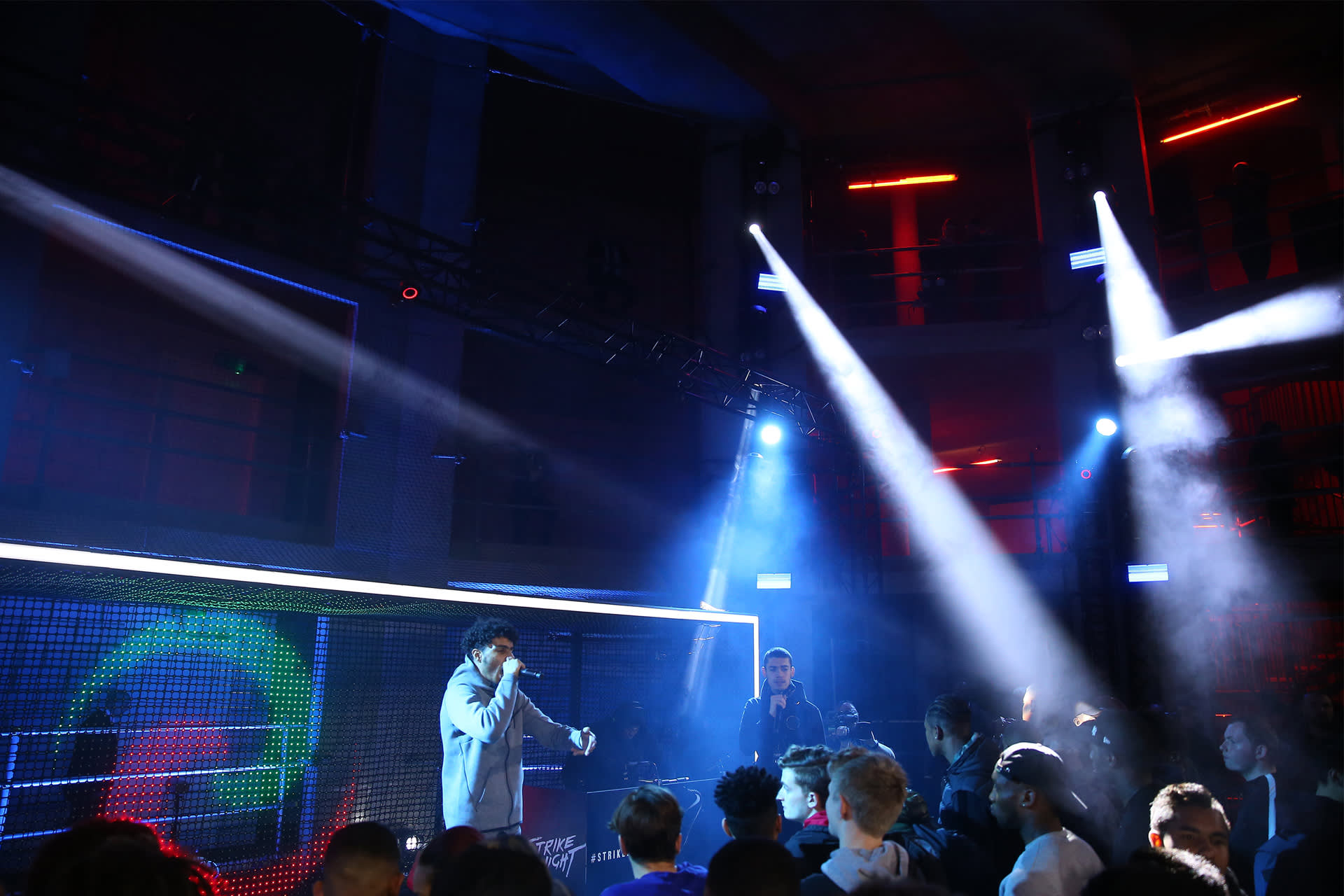 A performer sings on stage under bright spotlights at a crowded nightclub, with people in the audience looking toward the stage. The venue has colorful lighting and an elevated balcony.