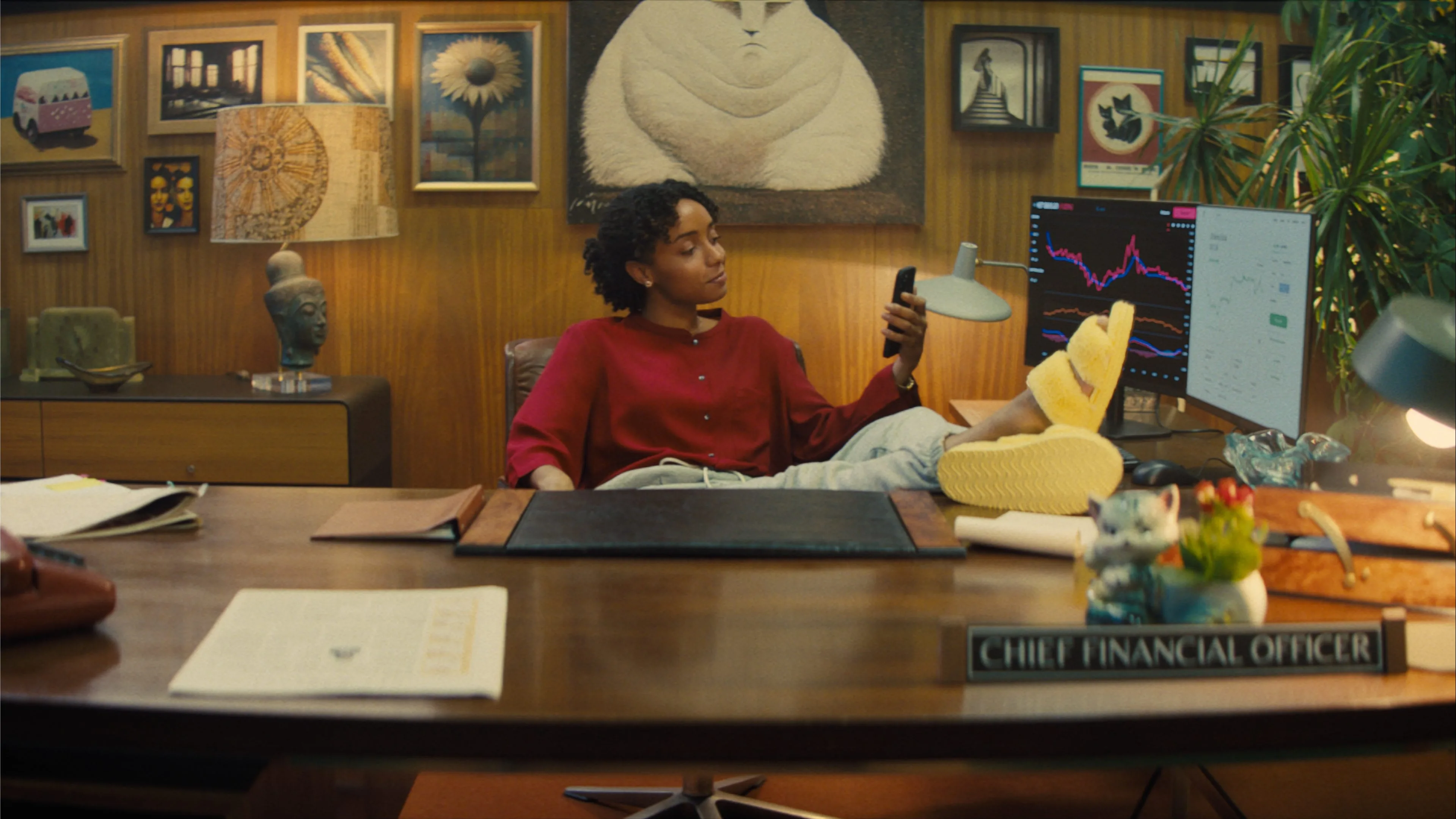A person in a red shirt and yellow slippers relaxes with feet on a desk in an office labeled Chief Financial Officer, looking at a phone. Behind them are art, books, and finance charts on a monitor.
