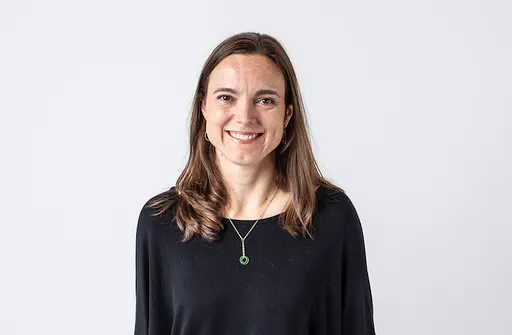 A woman with long brown hair, wearing a black top and a green pendant necklace, smiles at the camera against a plain white background.