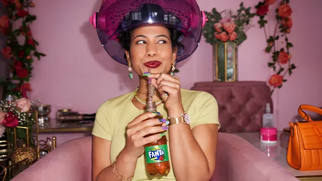 A woman sits under a hair dryer in a salon, smiling as she sips a Fanta drink through a straw. She wears a yellow shirt, statement earrings, and is surrounded by pink decor and flowers.