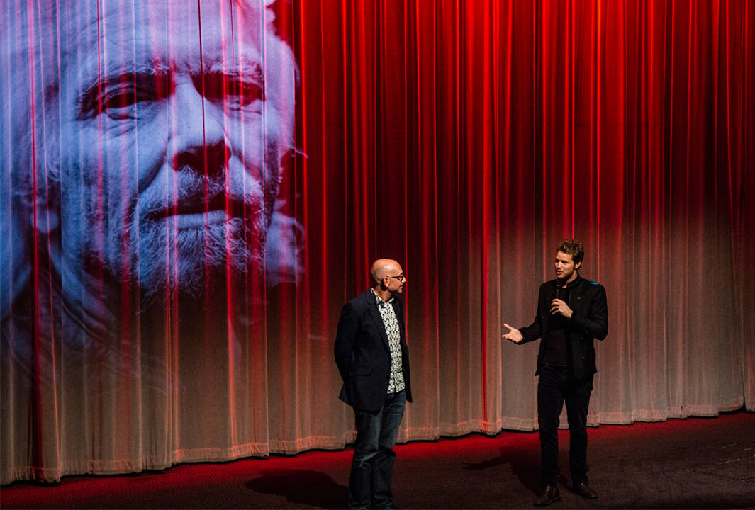 Two men stand and speak on stage in front of a large red curtain with a black-and-white projected image of an older mans face on it.