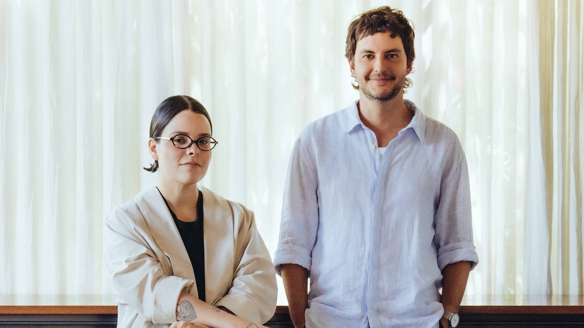 Two people stand indoors in front of sheer curtains. The person on the left wears glasses, a beige blazer, and has dark hair pulled back. The person on the right has short hair, a light blue shirt, and is smiling.