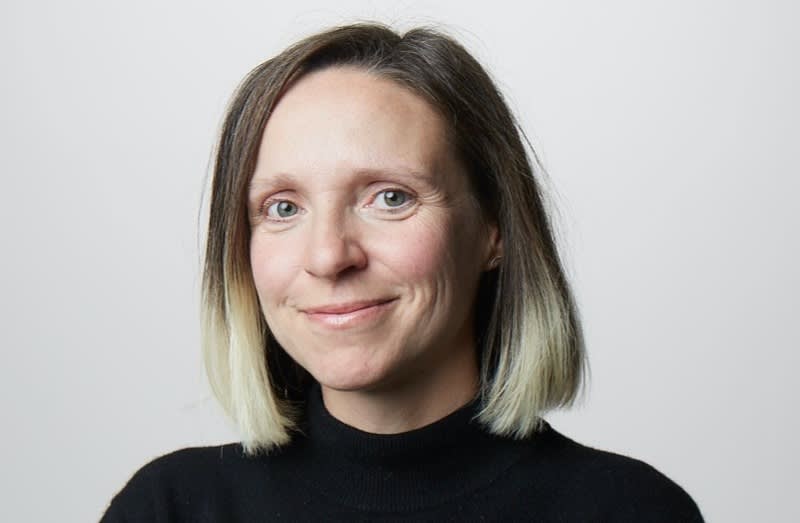 A woman with shoulder-length straight hair, dark with light blonde tips, wearing a black turtleneck, smiles softly at the camera against a plain light gray background.