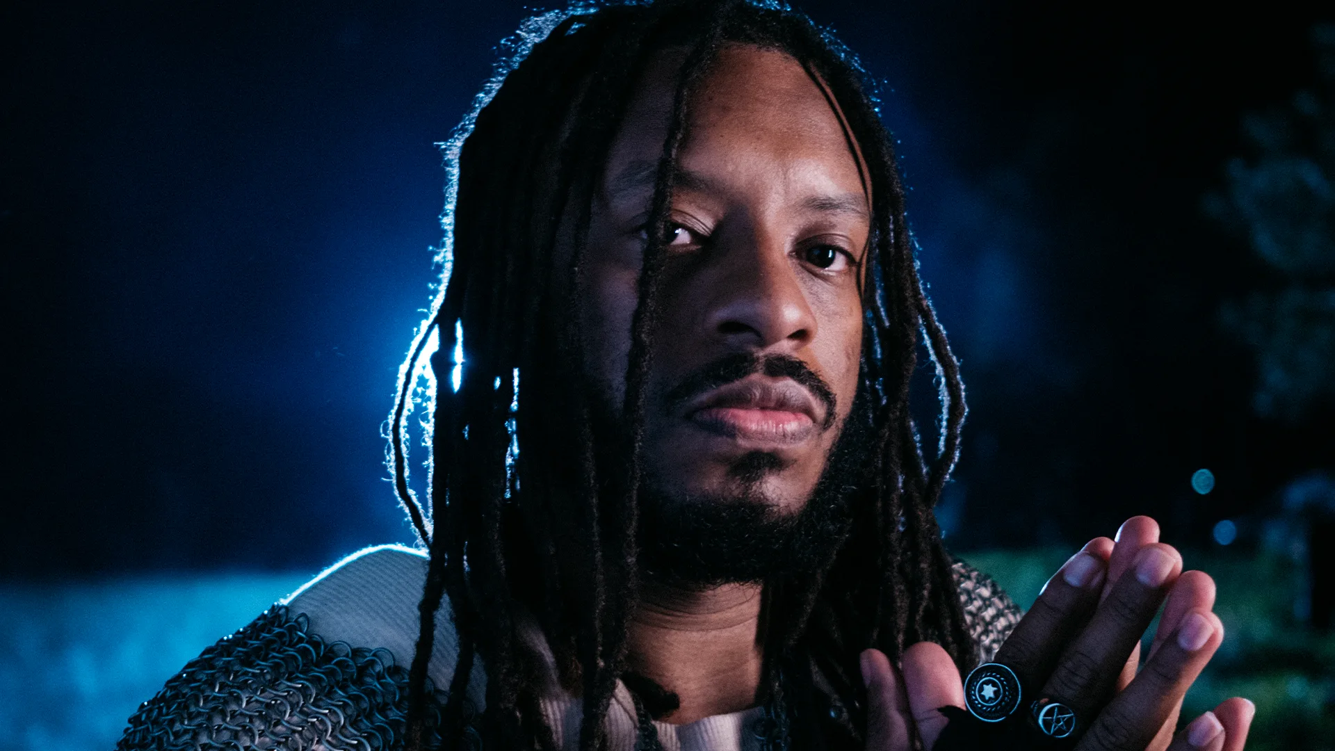 A person with long dreadlocks and a beard faces the camera at night, illuminated by blue light. They wear a chainmail garment and hold their hands together, showing a ring with a symbolic design.