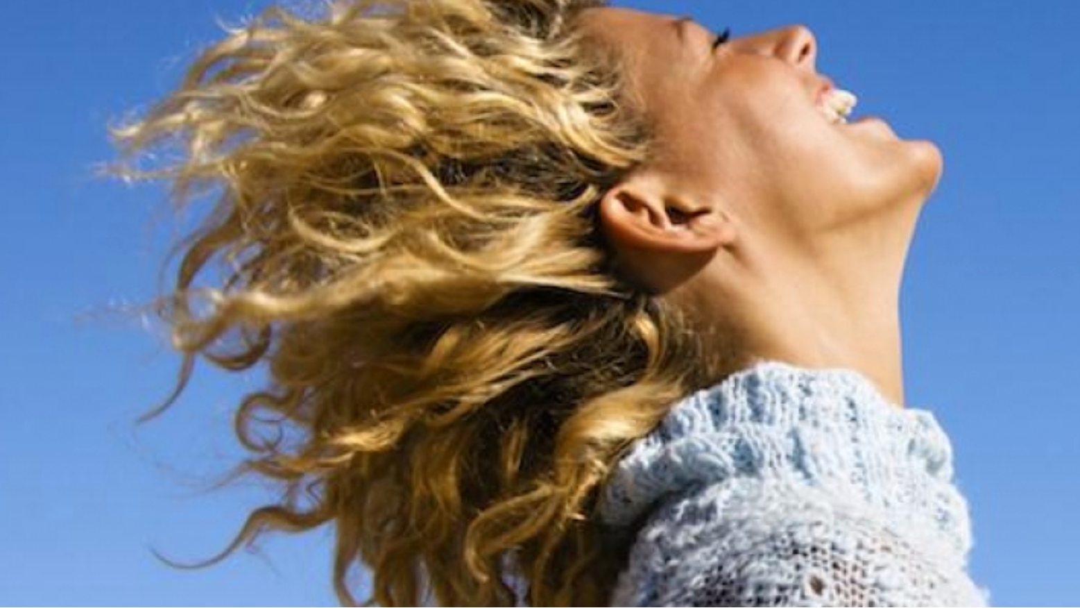 A person with curly blonde hair smiles joyfully, head tilted back, against a clear blue sky, wearing a light blue knitted sweater.