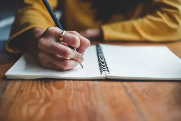 A person wearing a yellow long-sleeve top writes in a spiral notebook with a pencil on a wooden table. The focus is on their hand and the notebook, with the background blurred.