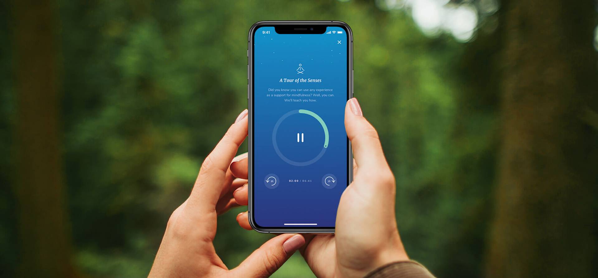 A person holds a smartphone outdoors displaying a meditation app screen with a pause button and a blue circular progress bar, against a blurred forest background.