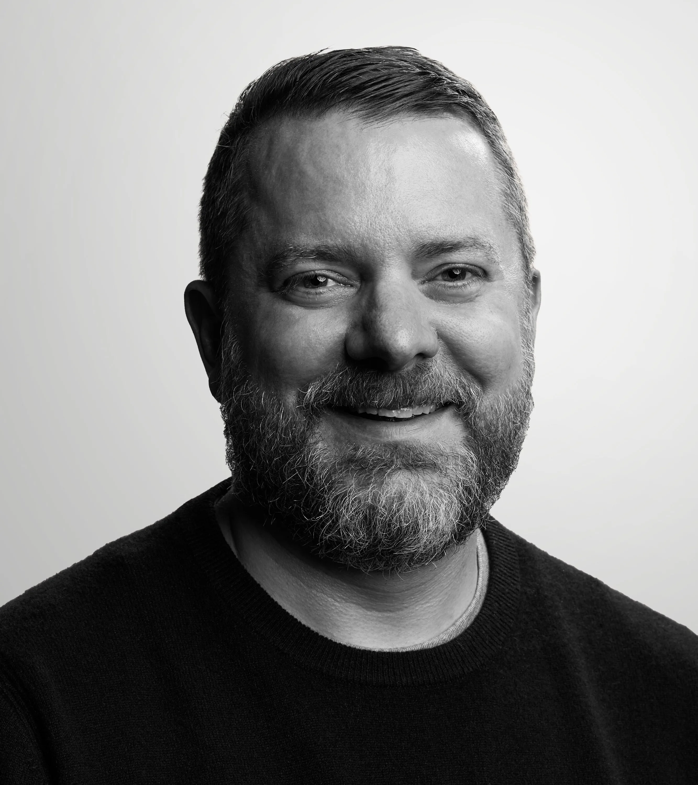 A bearded man with short hair, wearing a dark sweater, smiles while looking at the camera. The background is plain and light-colored. The photo is in black and white.