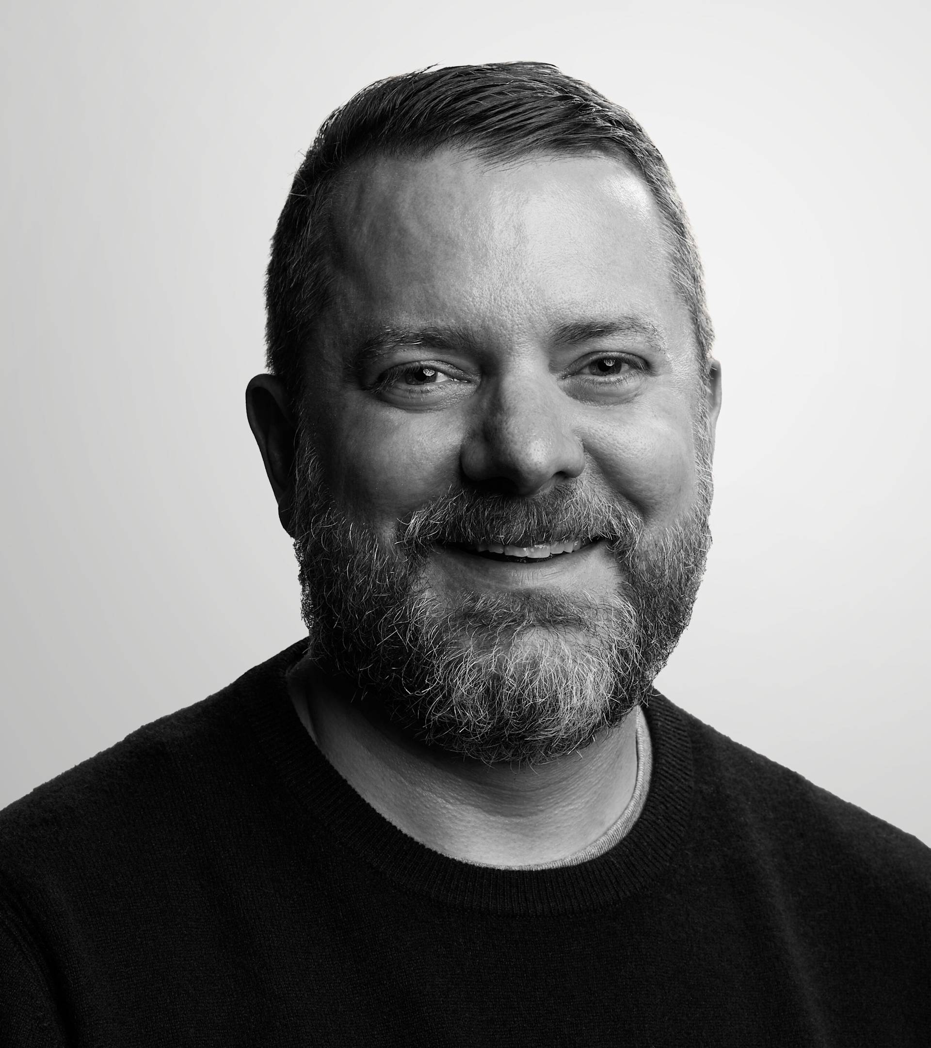 A bearded man with short hair, wearing a dark sweater, smiles while looking at the camera. The background is plain and light-colored. The photo is in black and white.