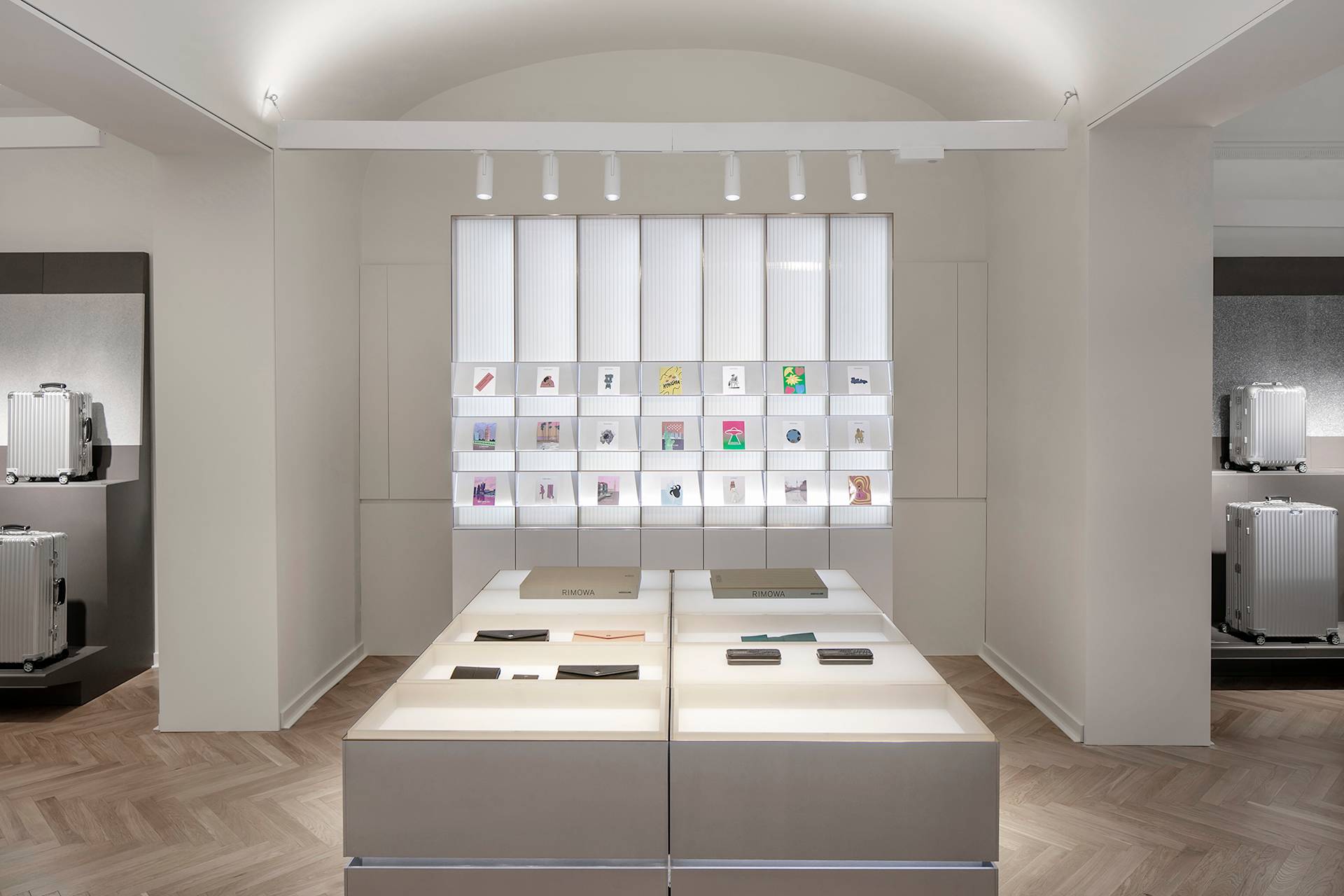 A modern, minimalist retail store interior with light wood floors, neutral walls, and illuminated display shelves holding various small items. Two display tables with accessories are centered in the foreground.