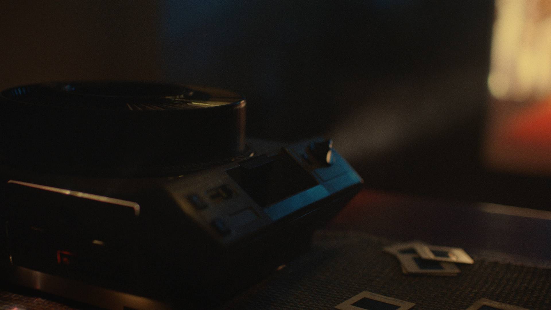 A close-up of a vintage slide projector on a table with several photo slides scattered nearby, illuminated by warm, low lighting.