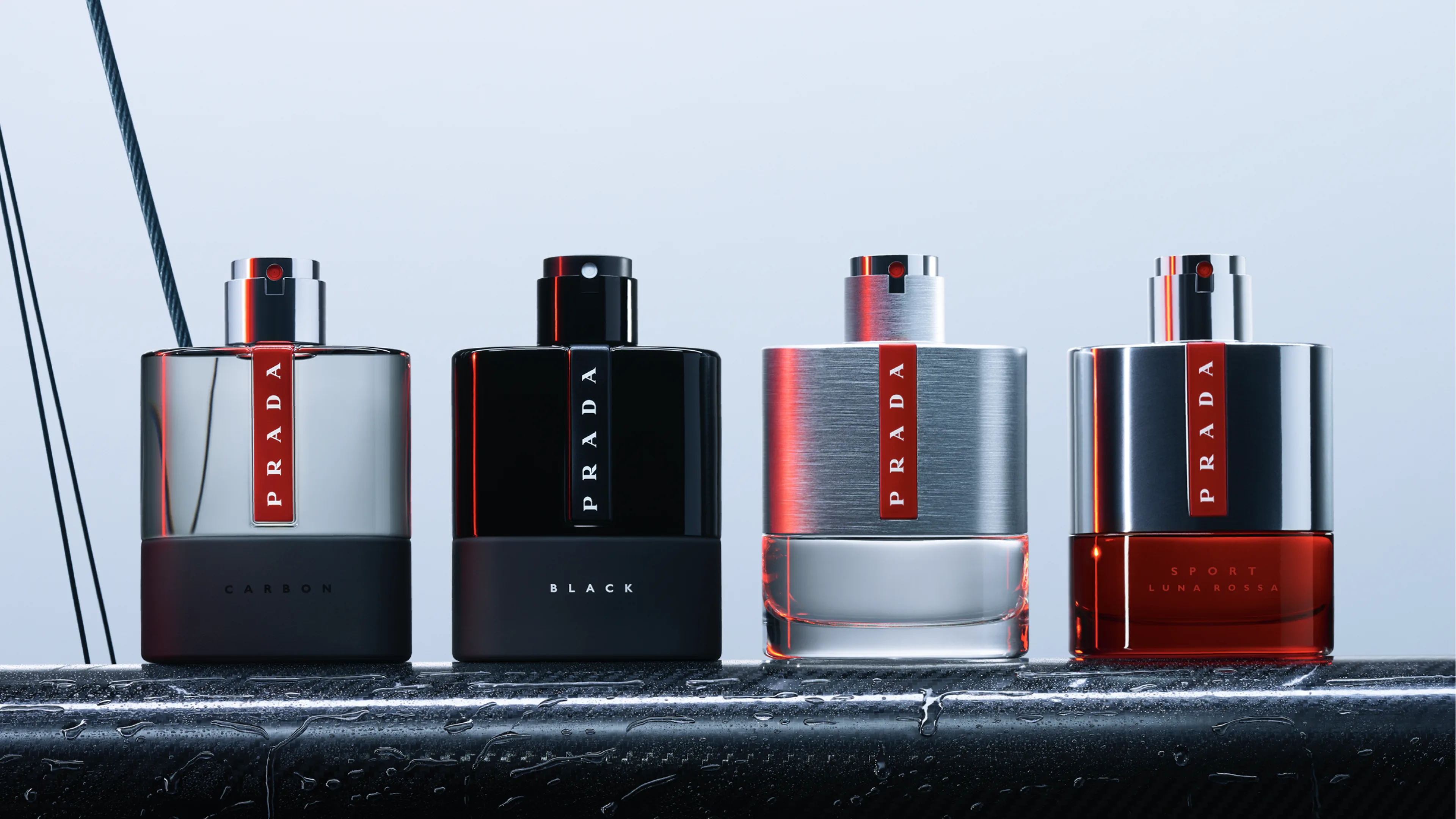 Four Prada perfume bottles—labeled Carbon, Black, and Sport Luna Rossa—stand side by side on a wet surface, against a light background with diagonal black lines. The bottles are sleek and modern in silver, black, and red tones.