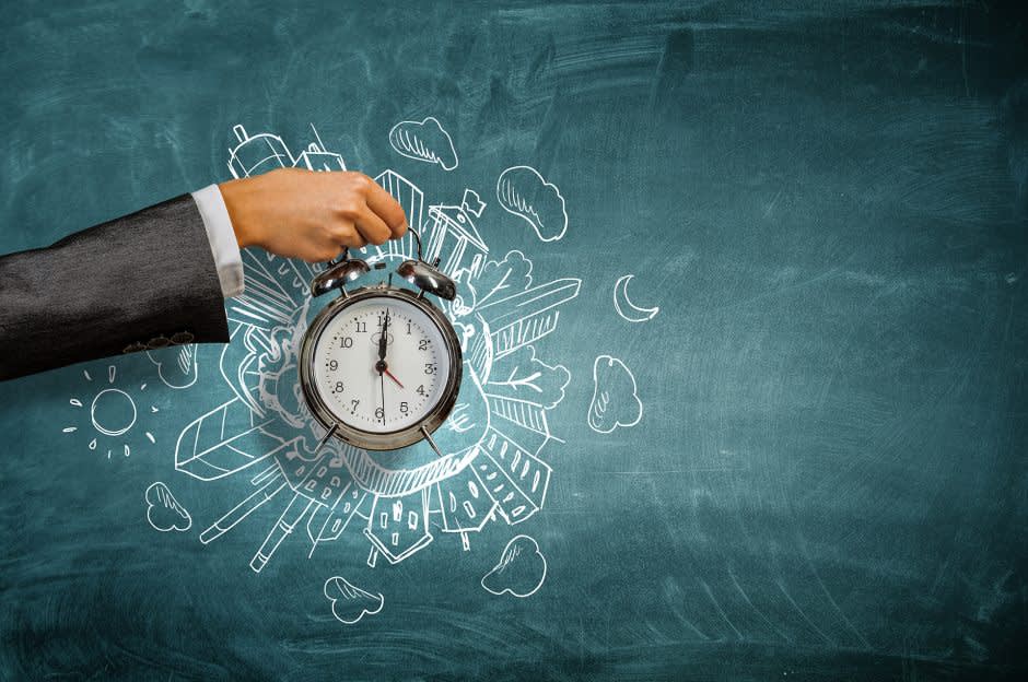 A person in a suit holds an alarm clock in front of a chalkboard drawing that features buildings, clouds, and the sun, symbolizing time management in a city or work environment.