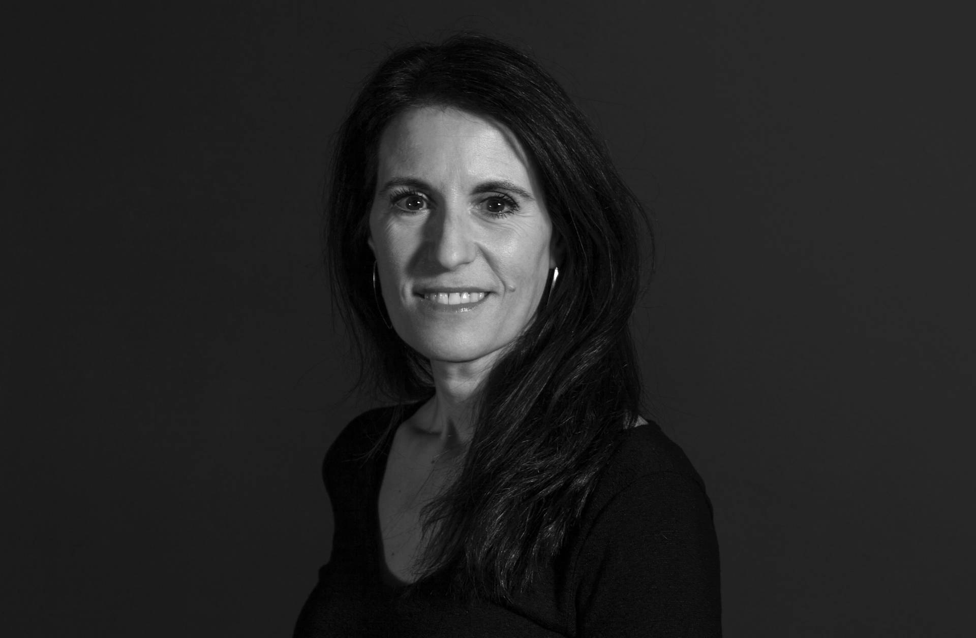 A woman with long dark hair and hoop earrings smiles slightly, wearing a black top, against a plain dark background in a black and white portrait.