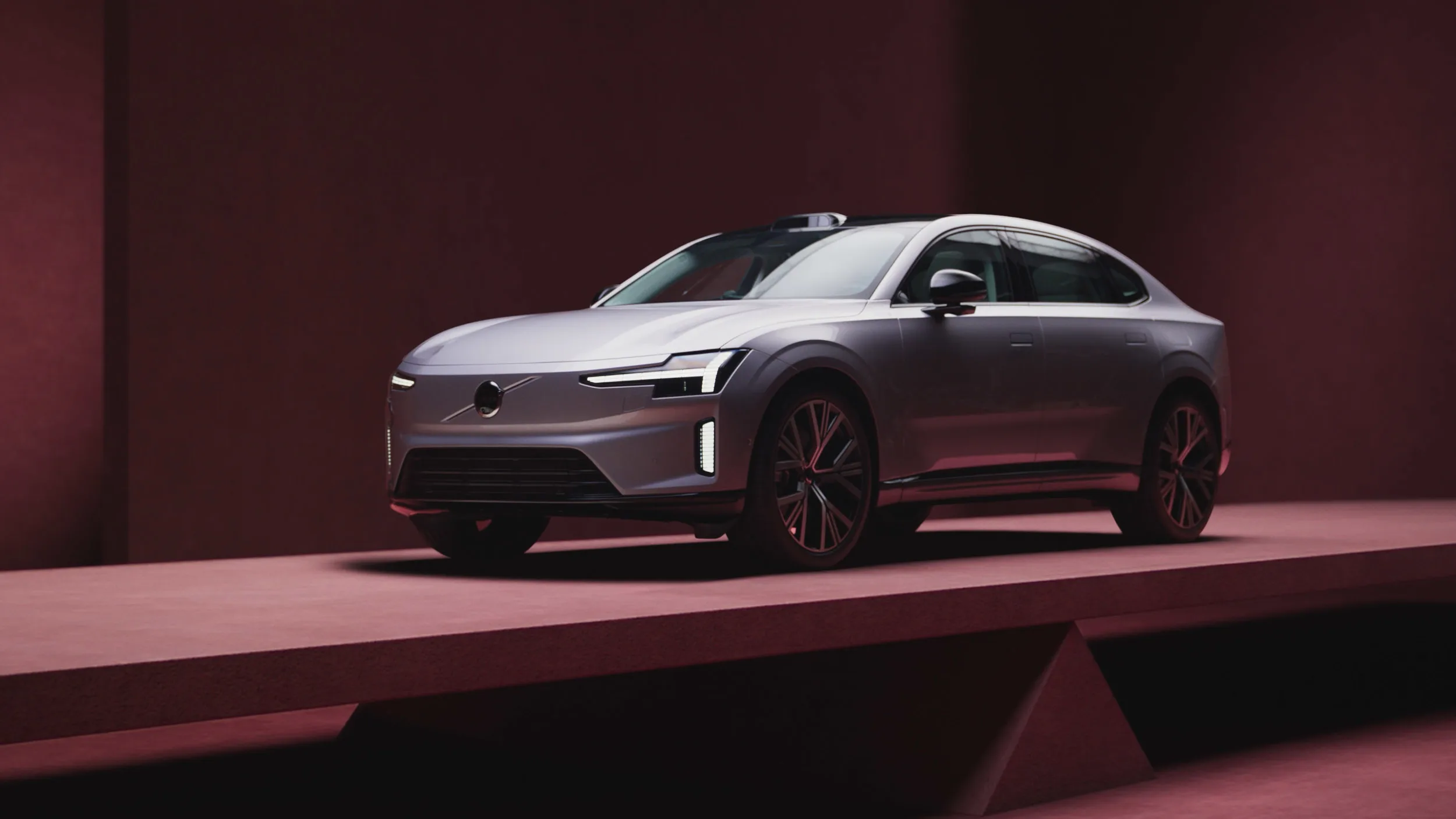 A sleek, silver modern sedan displayed on a geometric platform in a room with dark red walls, under soft lighting, highlighting its sharp lines and stylish design.