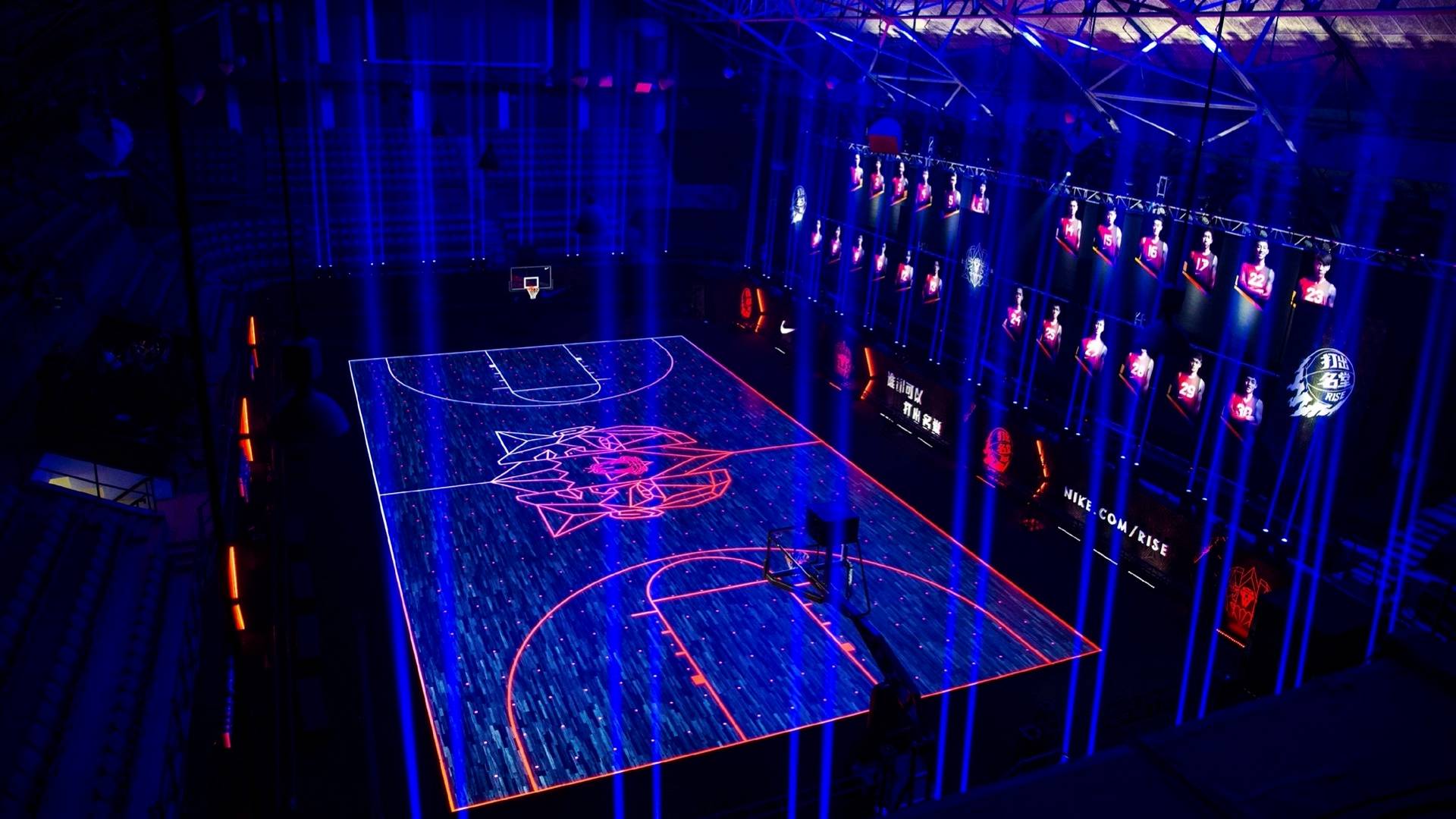A basketball court illuminated with vibrant blue and pink neon lights, featuring a bold center-court logo. Digital screens and spotlights surround the court, creating a futuristic, high-tech atmosphere in a dimly lit arena.