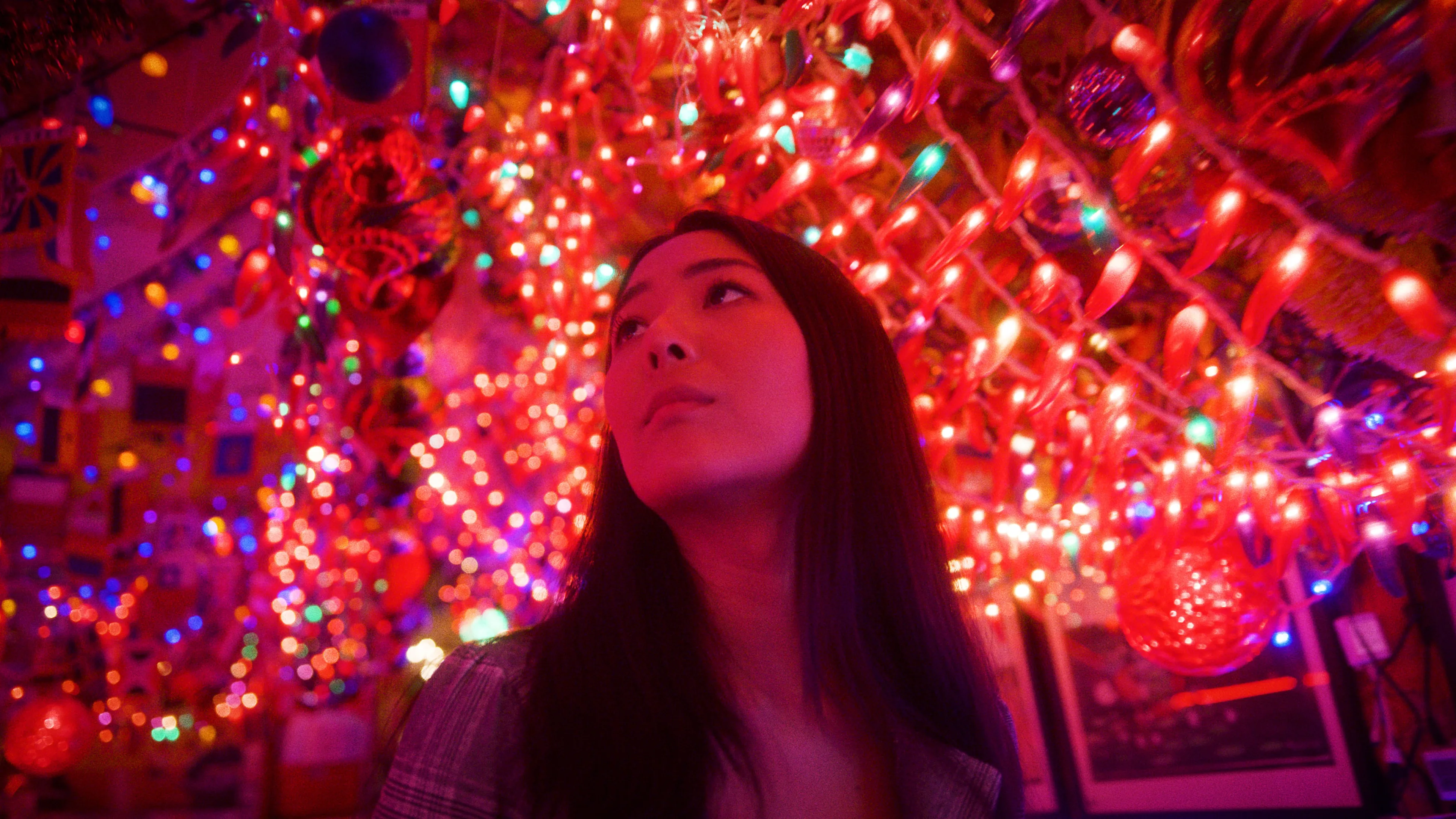 A woman with long dark hair looks upward, surrounded by vibrant, colorful Christmas lights and ornaments that fill the background with a festive, glowing atmosphere.