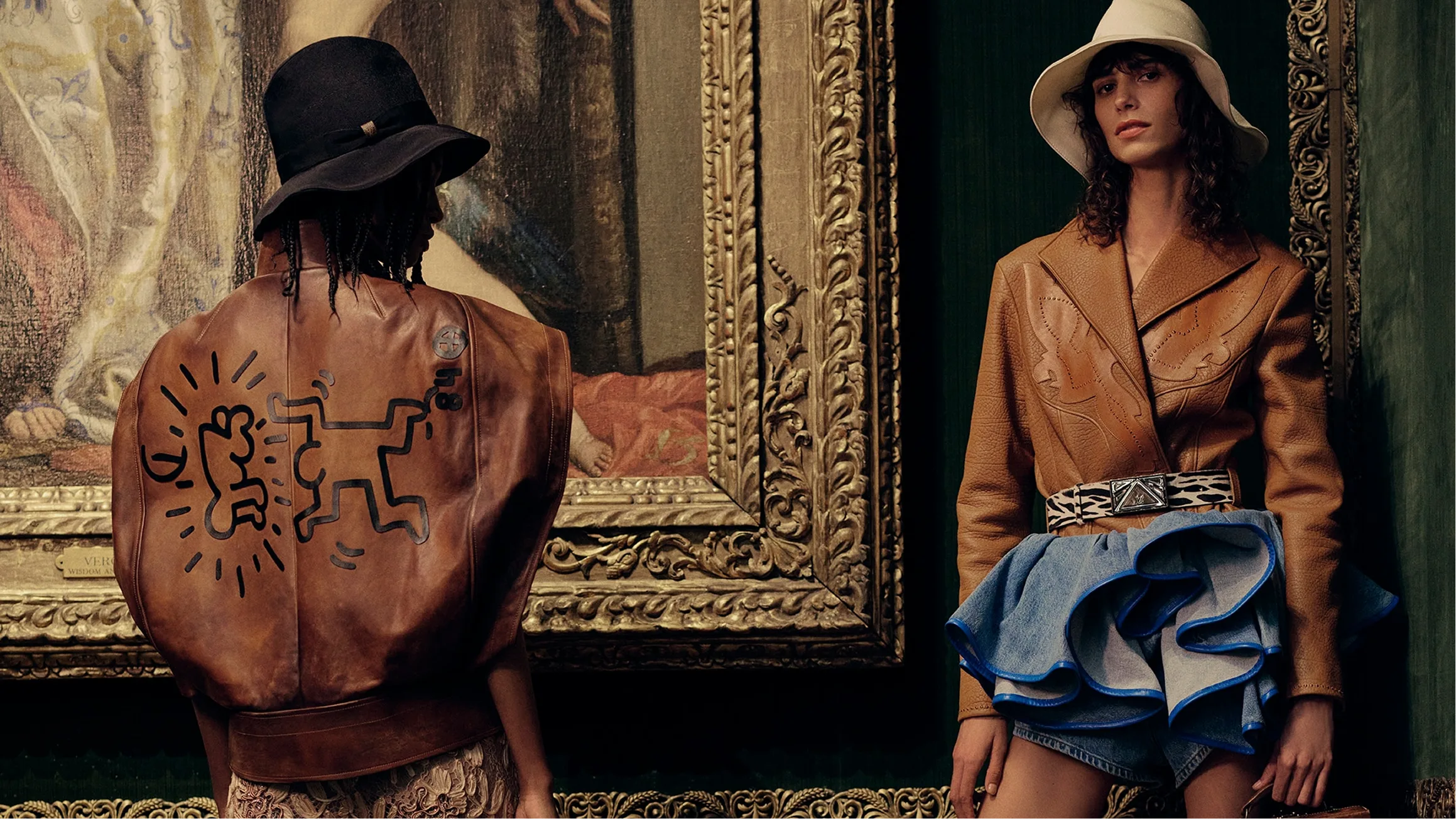 Two models stand in front of an ornate framed painting, one wearing a brown leather jacket with a black graphic print and the other wearing a tan blazer with a ruffled denim skirt.