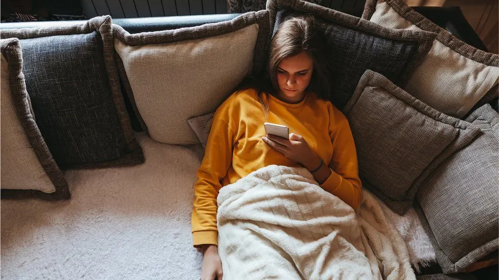 A young person in a yellow sweatshirt lies on a couch covered with a blanket and many pillows, looking at a smartphone.