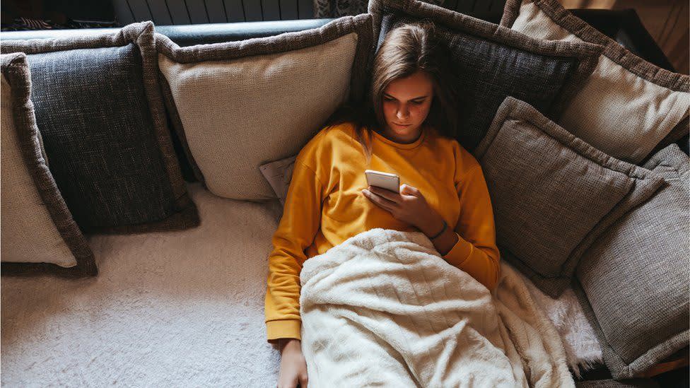 A young person in a yellow sweatshirt lies on a couch covered with a blanket and many pillows, looking at a smartphone.