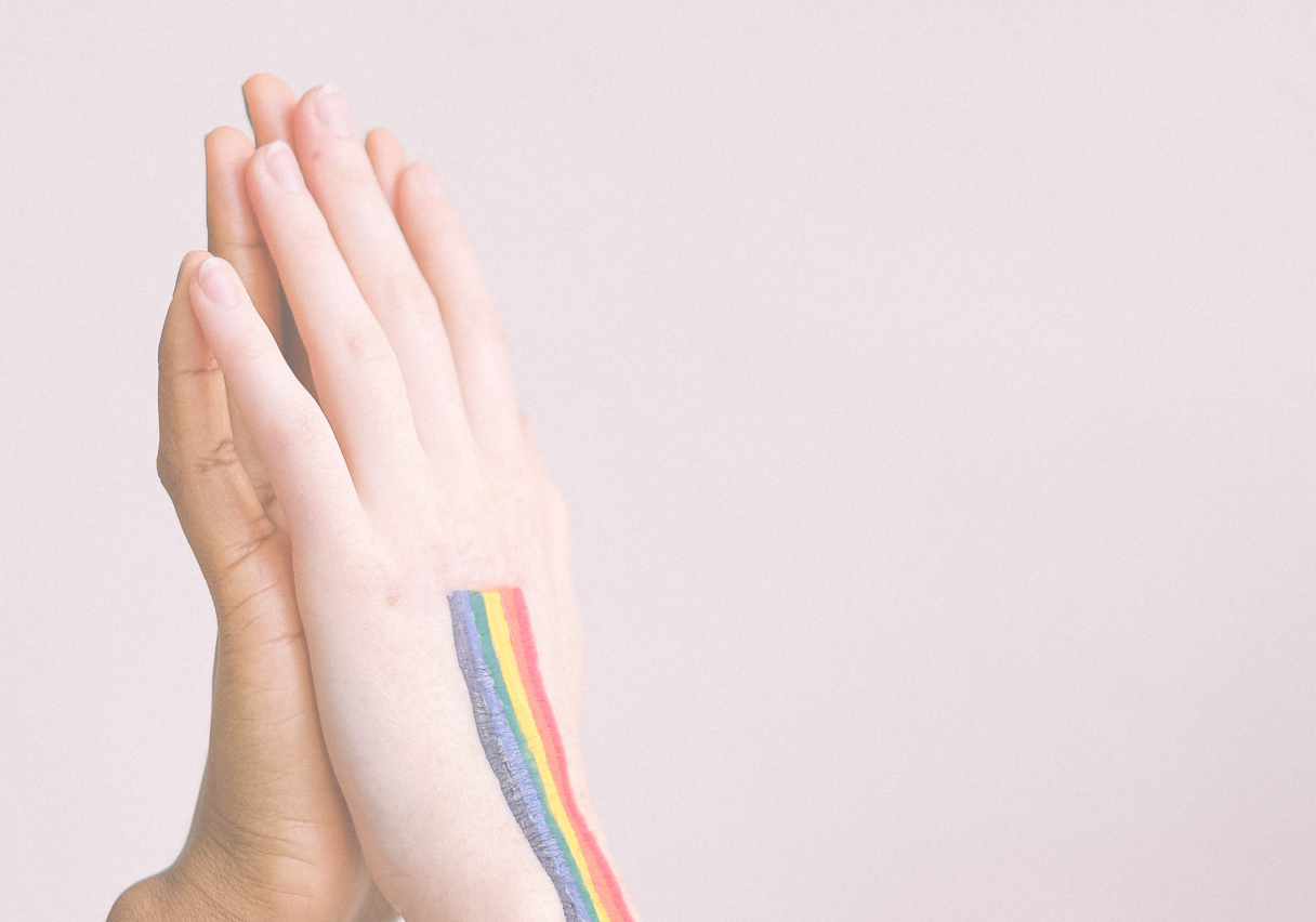 Two hands pressed together in a high-five gesture. One hand has a rainbow stripe painted on the wrist. The background is plain and light-colored.
