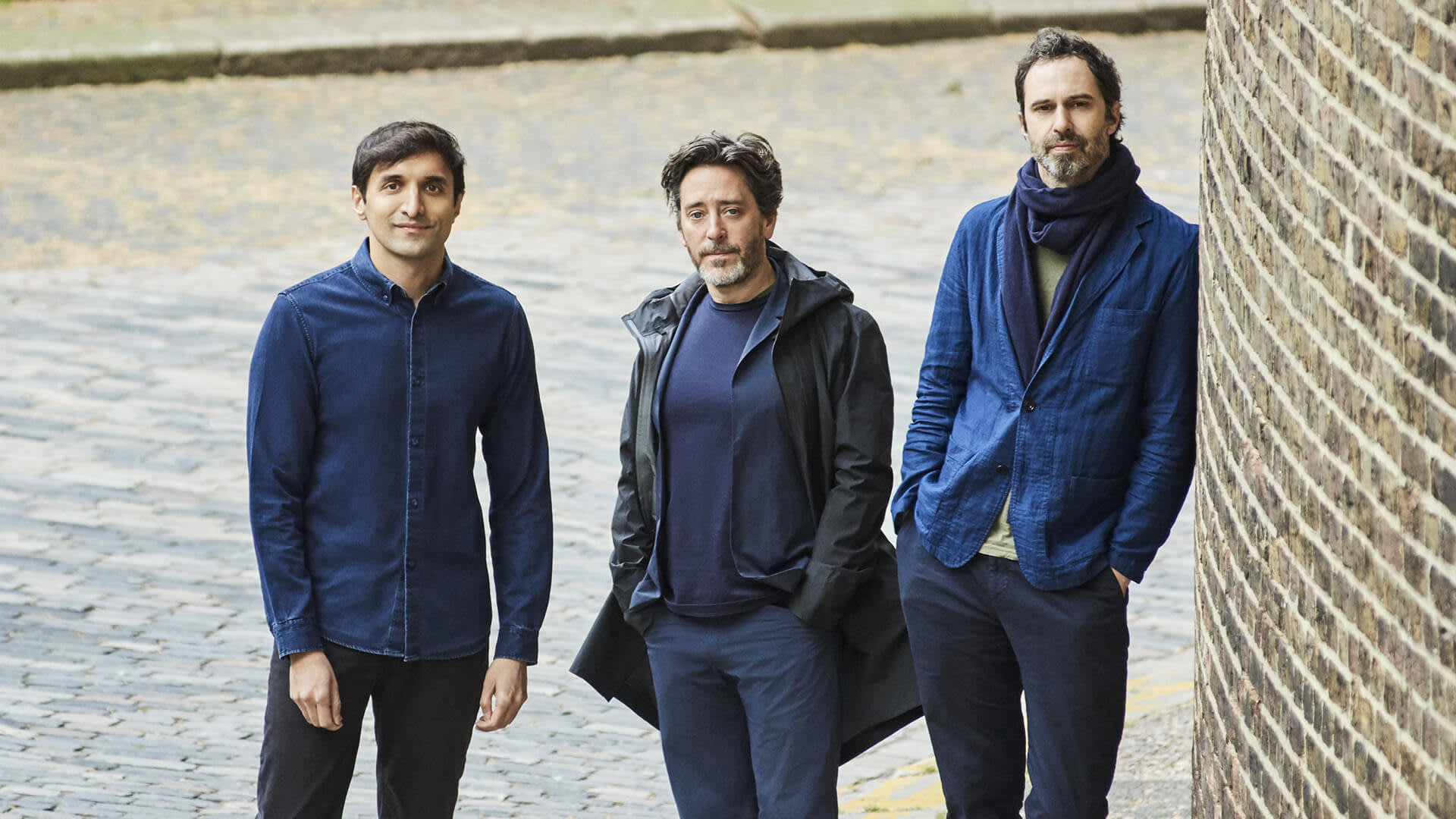 Three men stand outdoors on a cobblestone street beside a brick wall, wearing casual blue and dark-colored clothes. One leans against the wall, while the other two stand with hands in pockets, looking at the camera.