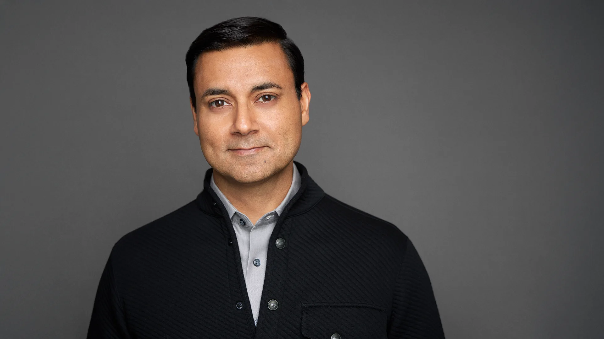 A man with short dark hair, wearing a gray collared shirt and a dark textured jacket, stands in front of a plain gray background, looking at the camera with a neutral expression.