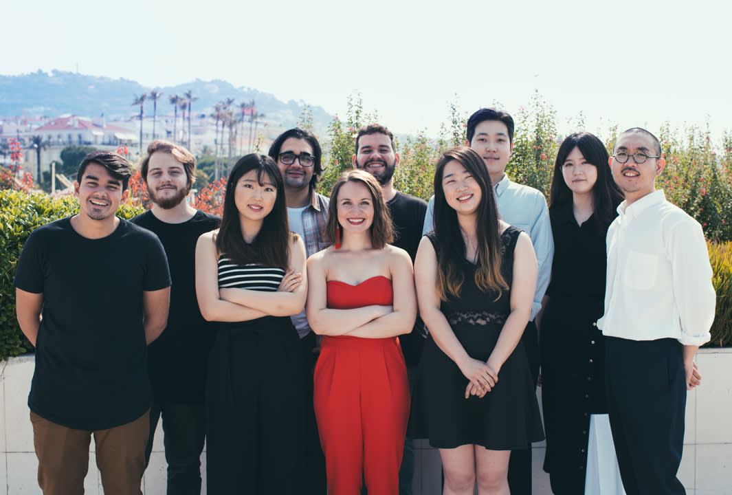 A group of ten people stand outside, smiling at the camera. They are dressed in casual and semi-formal clothes, with greenery and a cityscape visible in the background.