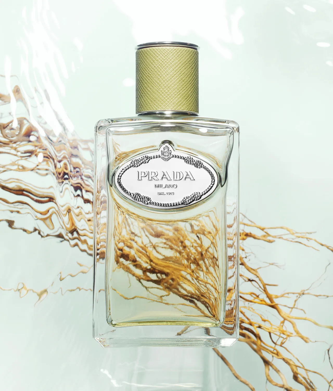 A Prada perfume bottle with a textured gold cap and an ornate label floats above swirling golden-brown strands in clear water, creating a luxurious and refreshing visual effect.
