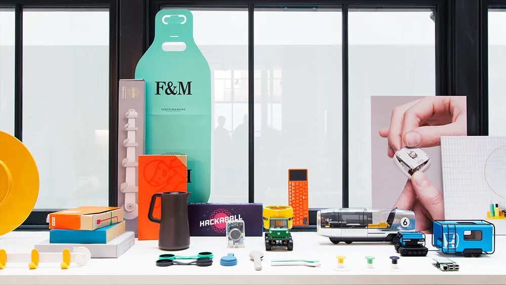 A colorful display of modern gadgets, toys, and boxes is arranged on a table in front of large windows. Items include a green F&M cutout, a calculator, toy vehicles, tech devices, and packaging.