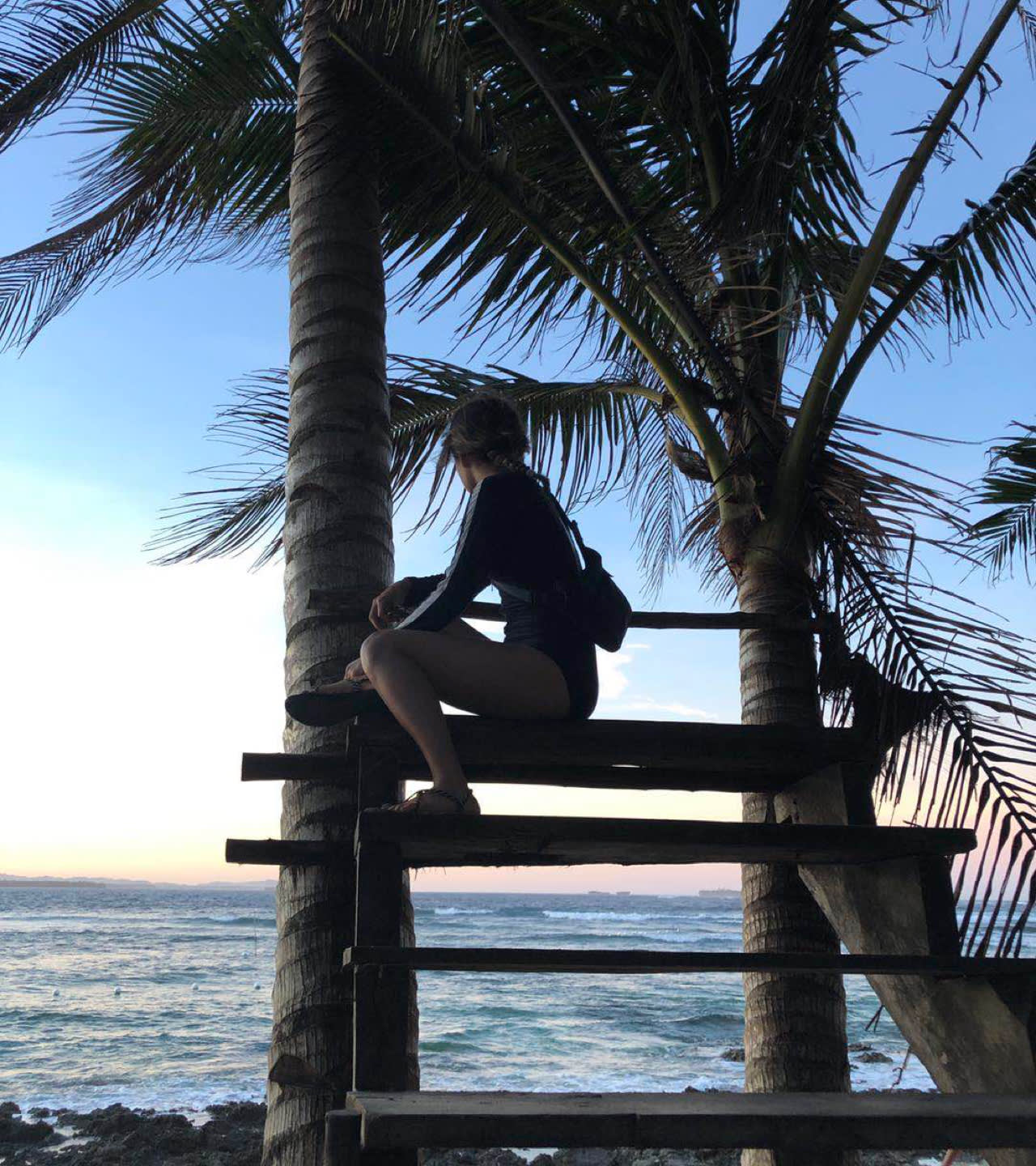 A person sits on a wooden platform built between palm trees, overlooking the ocean at sunset. The sky is a mix of blue and orange, and waves crash gently on the rocky shore below.