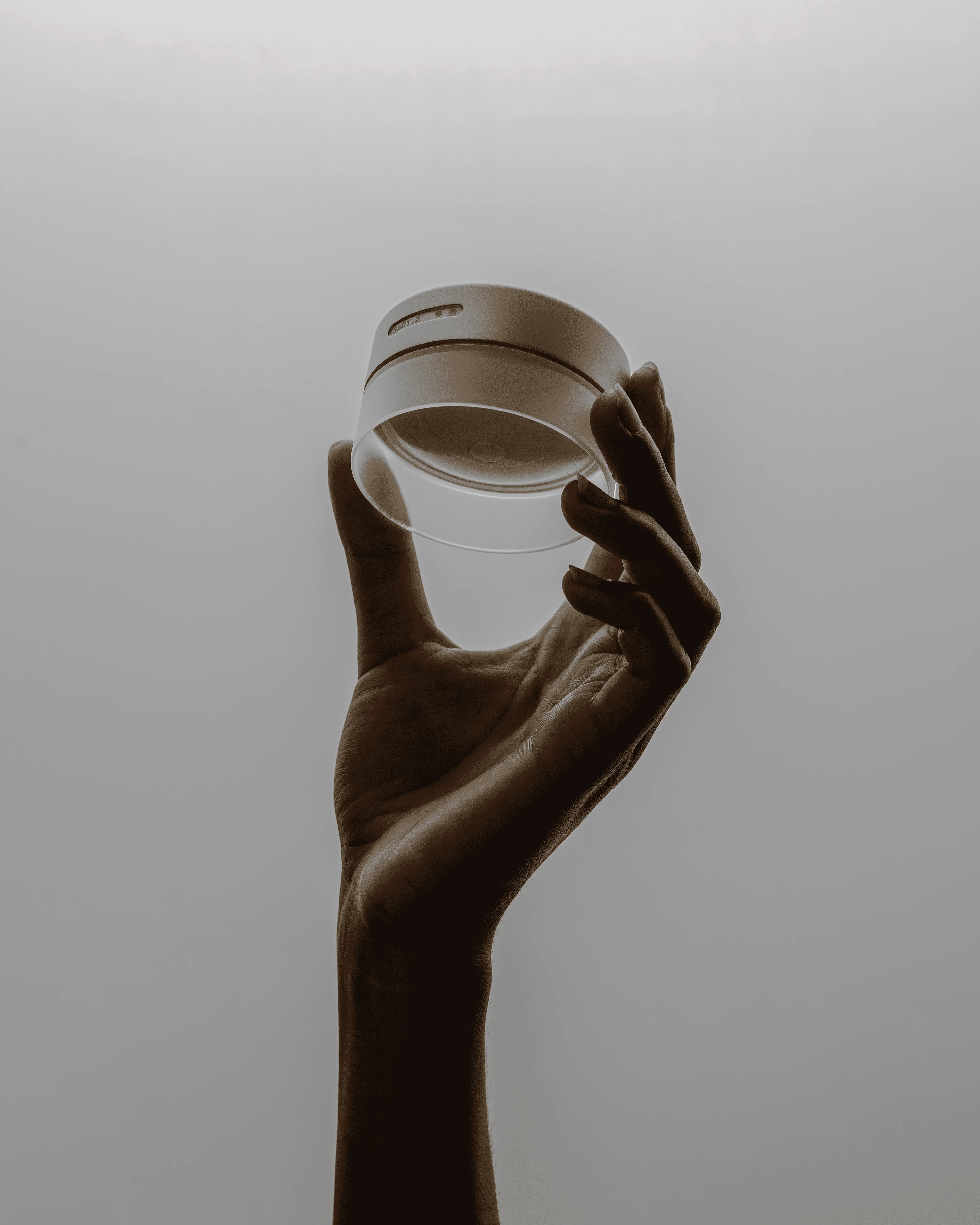 A hand holds up a round, transparent object, possibly a glass or lens, against a plain, softly lit background with a gradient from light to dark.