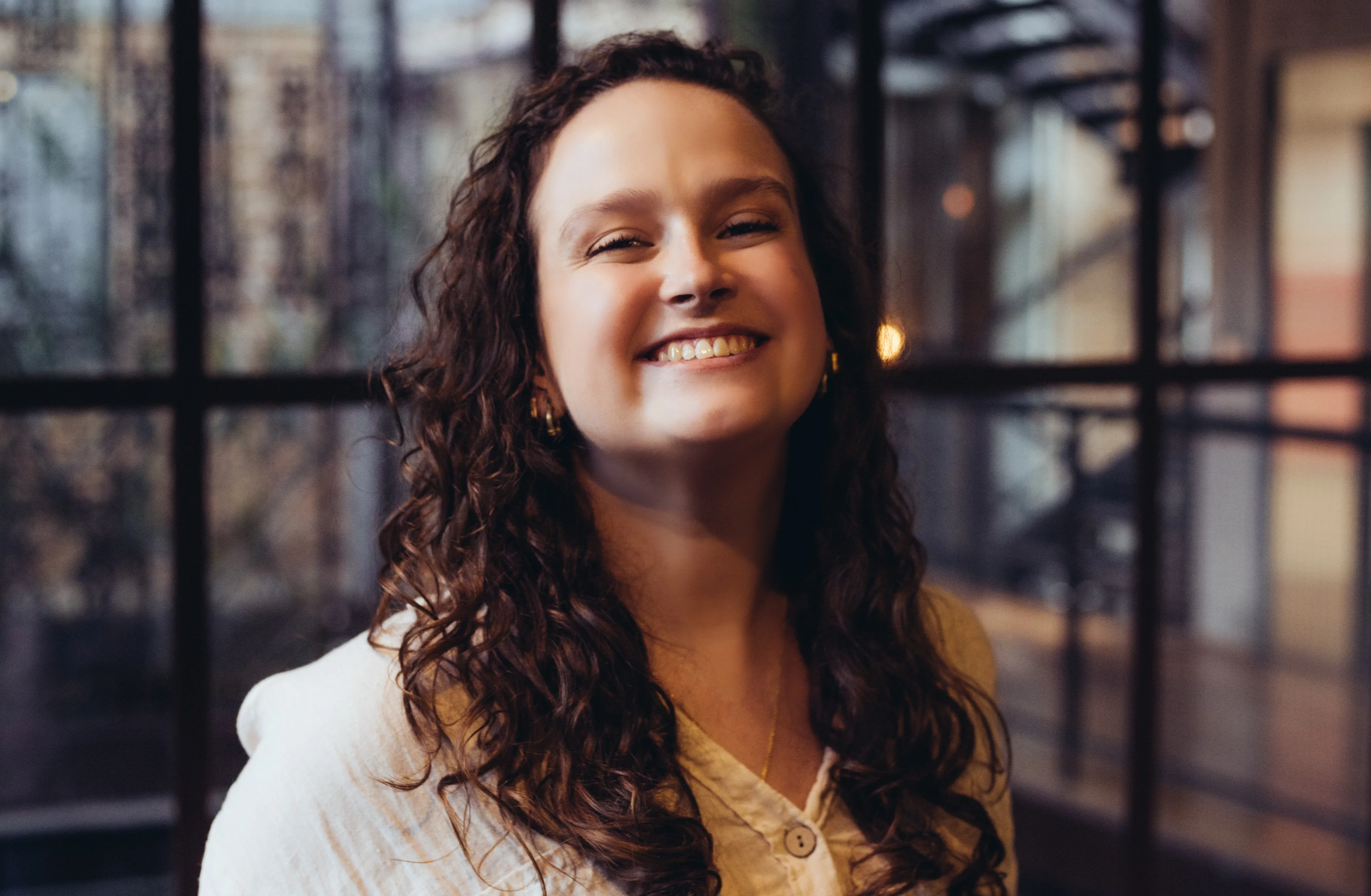 A woman with long curly brown hair, wearing a white shirt, smiles brightly while standing indoors in front of large windows with blurred surroundings.