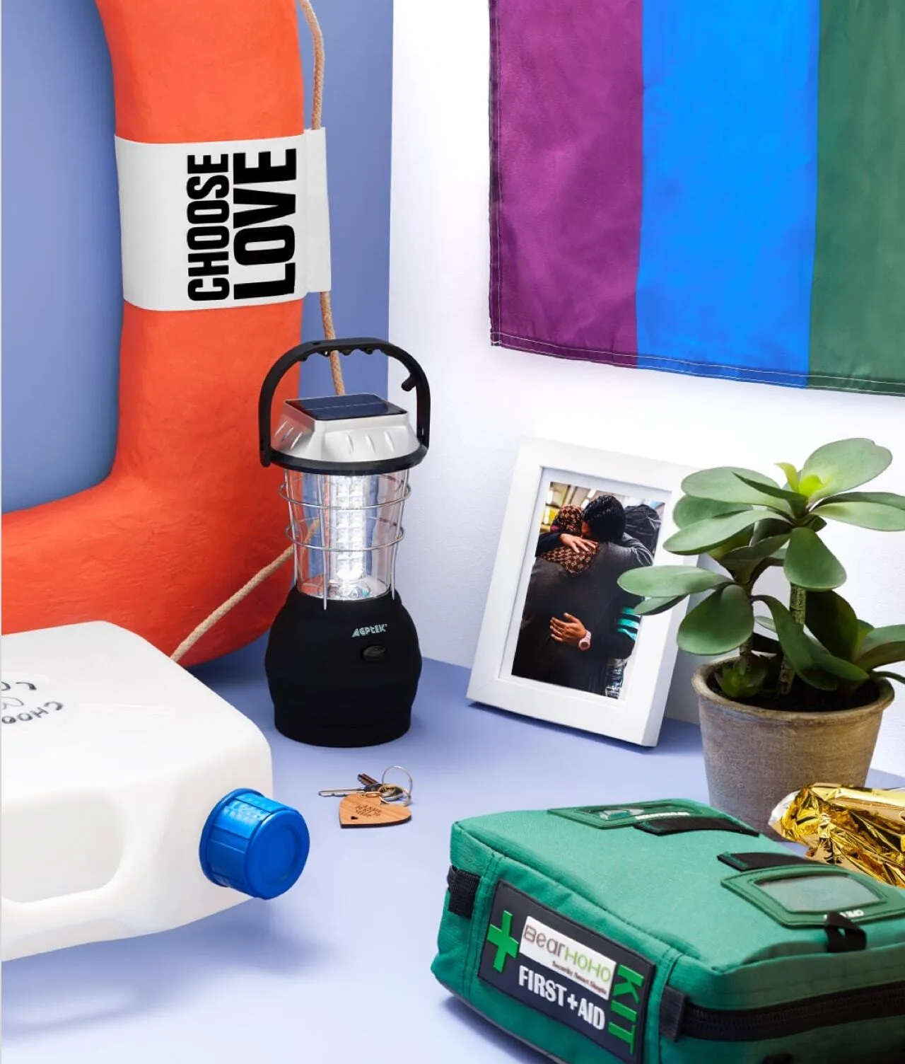 A life preserver labeled “CHOOSE LOVE,” a lantern, a first aid kit, a jug of water, keys, a potted plant, a framed photo of two people hugging, and a rainbow-striped flag on a light blue surface.