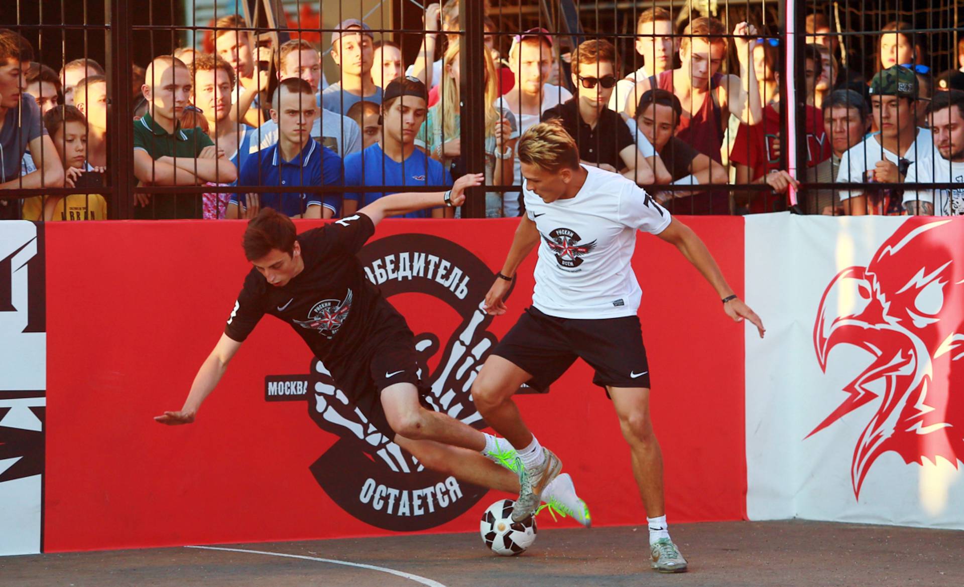 Two young men compete for the soccer ball on a small outdoor court, with one player in black stumbling as the other in white controls the ball. Spectators watch behind a fence and red and white banners.