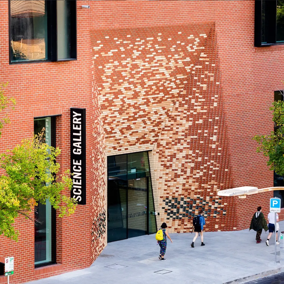 A modern brick building with a unique entrance where bricks are arranged in a pixelated, wave-like pattern. A black sign reads SCIENCE GALLERY. Several people walk on the sidewalk in front. Trees and a parking sign are nearby.