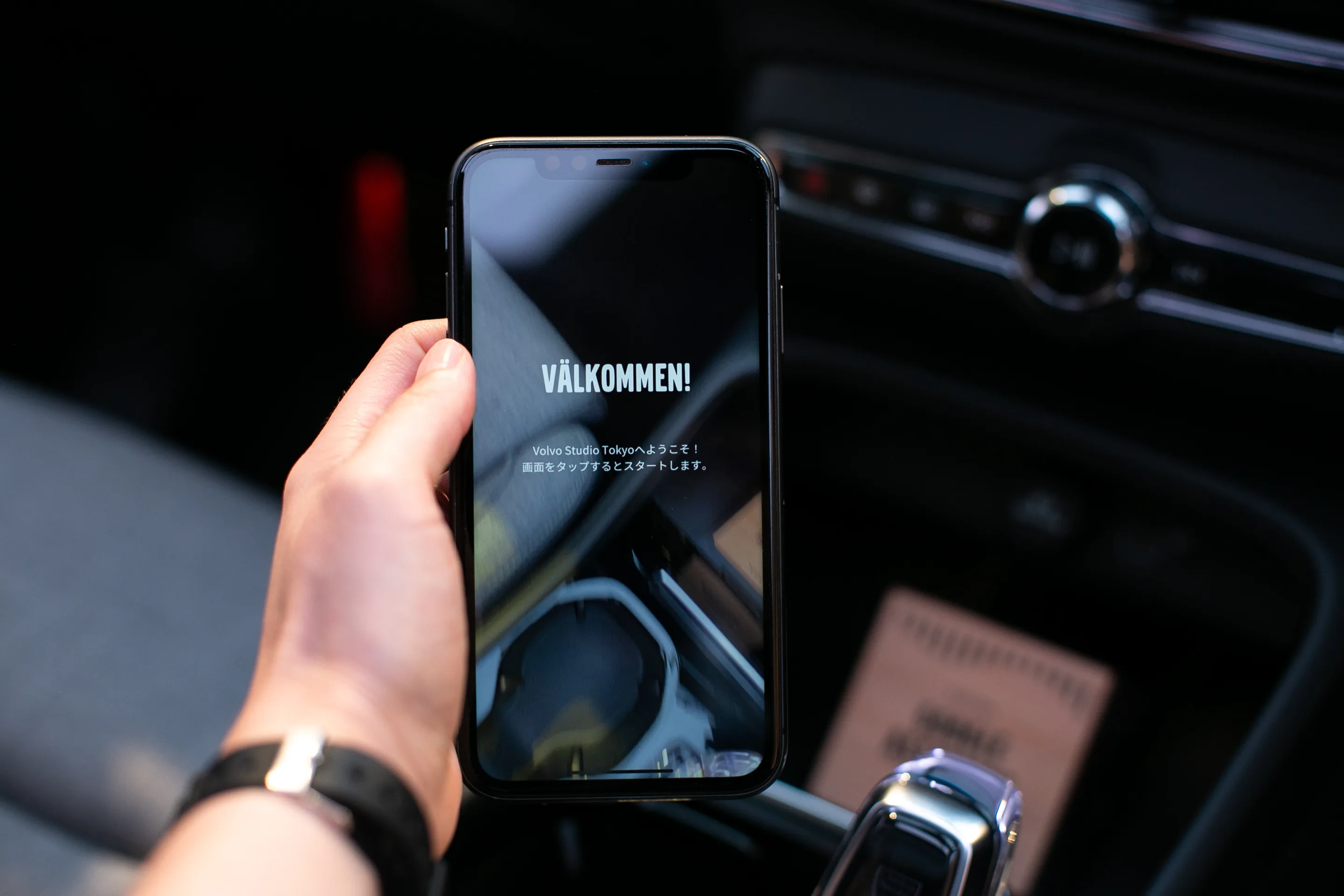 A hand holds a smartphone displaying a welcome screen with the word VÄLKOMMEN! in large text and smaller Japanese text below, inside a car with blurred interior details in the background.