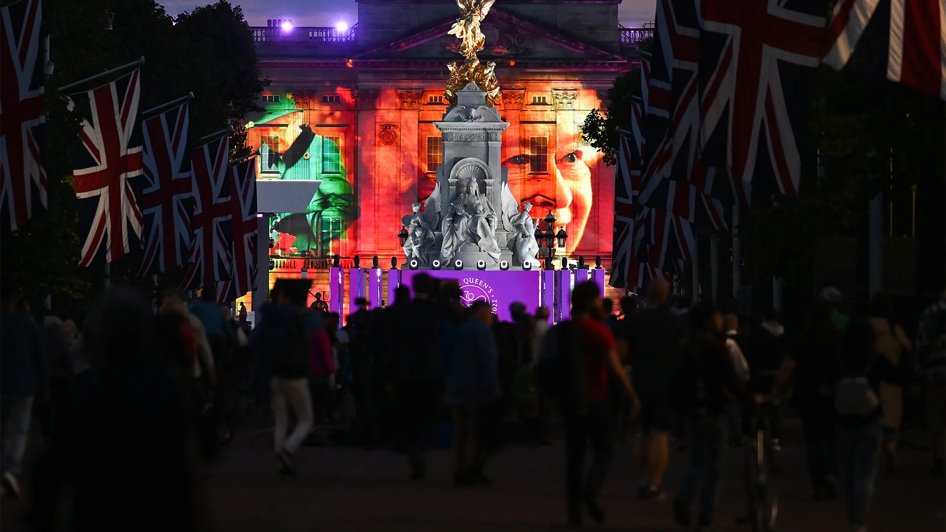 A crowd walks along a flag-lined street toward a brightly lit monument with the image of a smiling woman projected onto a building at night. Union Jack flags hang on both sides.