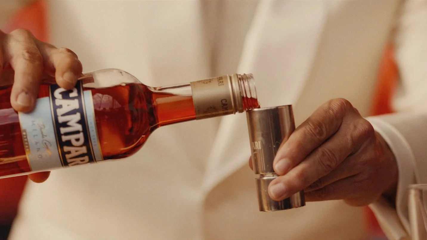 A person in a white suit pours Campari liqueur from a bottle into a metal jigger, preparing a drink.
