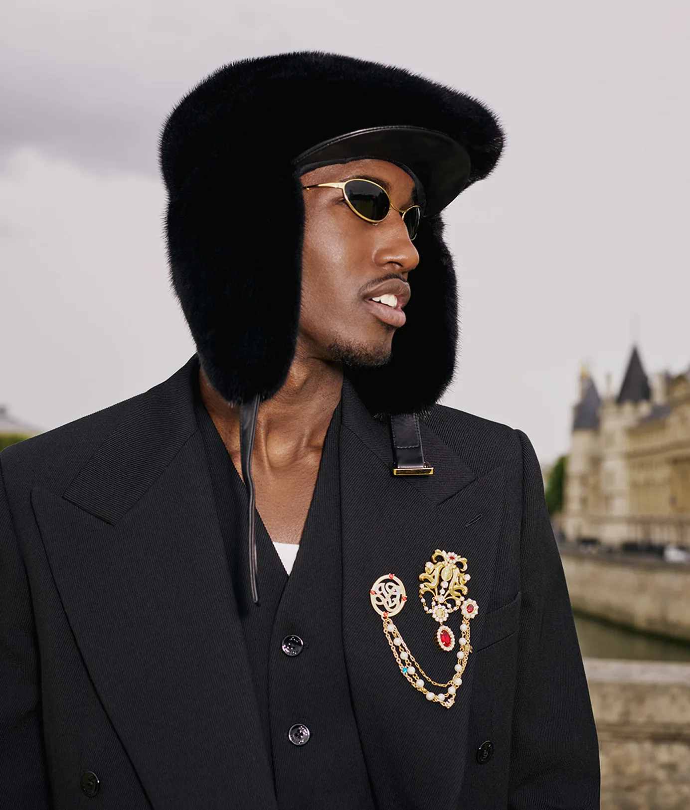 A man wearing a black fur ushanka hat, dark oval sunglasses, and a black suit jacket adorned with an ornate gold brooch featuring pearls and colorful jewels.